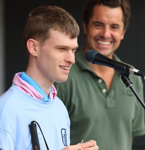 Tyler stands in front of a microphone smiling wearing a blue and pink shirt. A man stands behind him smiling wearing a green shirt.