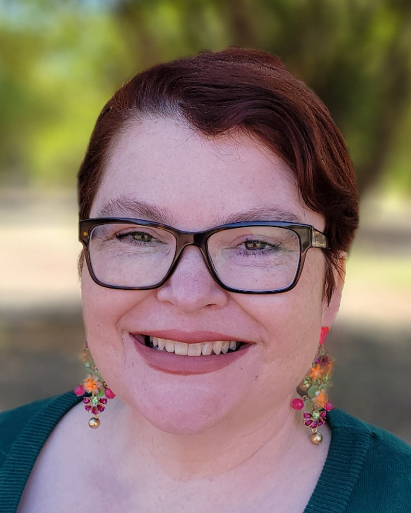 A woman with short, auburn-colored hair, glasses, and pale freckled skin smiles before a faded background of orange trees.