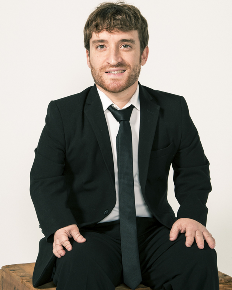 Nic Novicki a white male little person with a little facial hair, sits on a stool wearing a black suit, white shirt and skinny black tie.