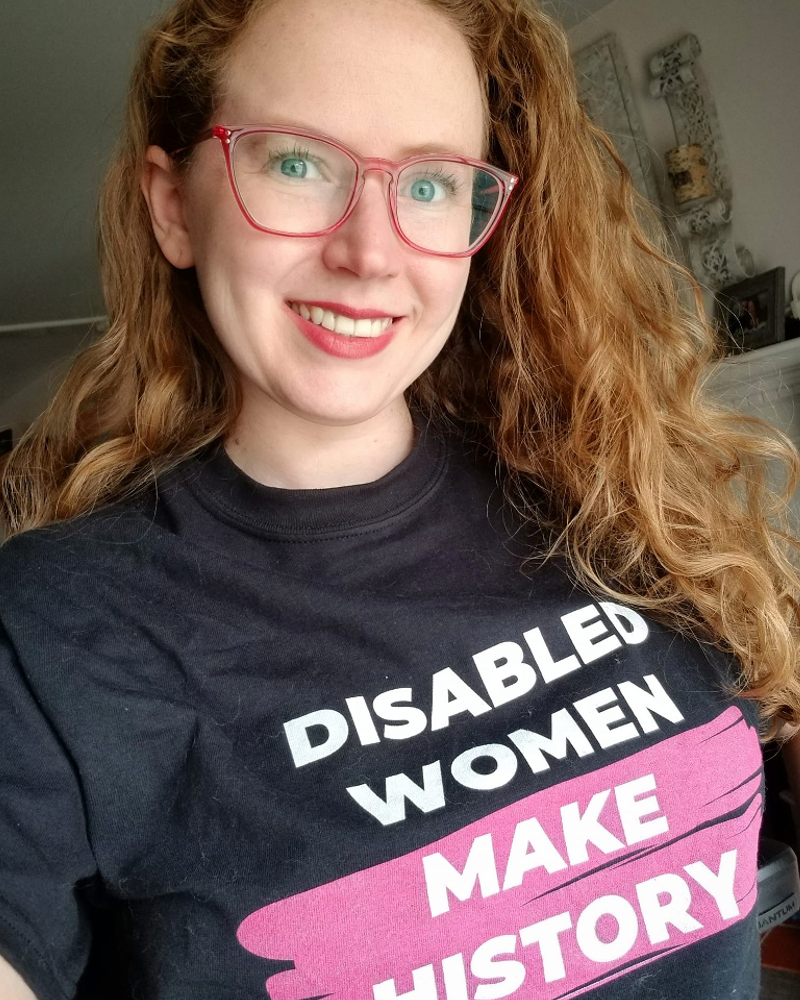 Stephanie Woodward, a pale woman with curly ginger hair and pink glasses, smiles while wearing a black shirt that reads 