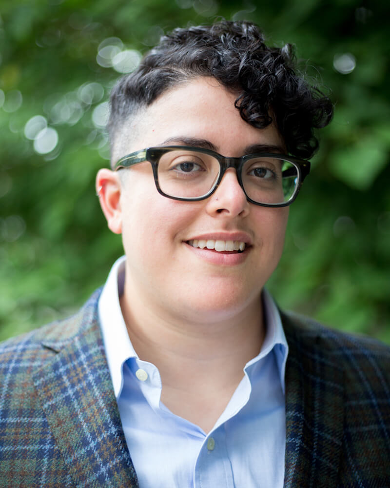 Aimi Hamraie, an olive-skinned Iranian person with short dark curly hair, smiles at the camera. They wear rectangular glasses and a blue button-up shirt. Behind them is a blurry green background