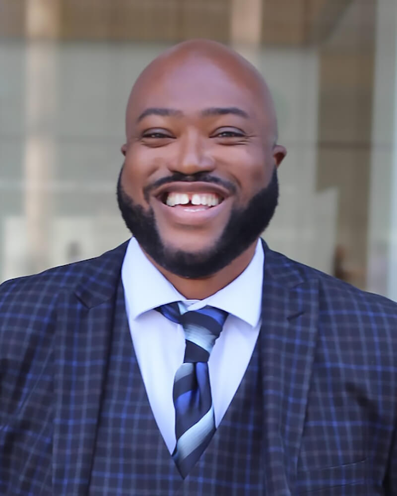 An African American man standing tall behind a building with huge glass windows; he's wearing a black and blue stripe suit with a blue dress shirt and dark blue tie he's bald and smiling big with his hands in his pockets.