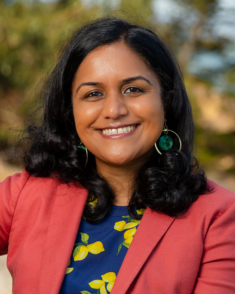 A Brown, South Asian female sitting outside with an orange blazer over a dark blue shirt with lemons.