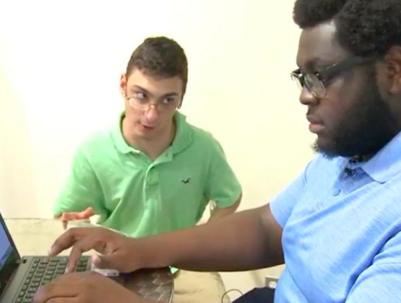 African American male peer mentor wearing a light blue shirt typing at a laptop with with white male mentee seated to his right.