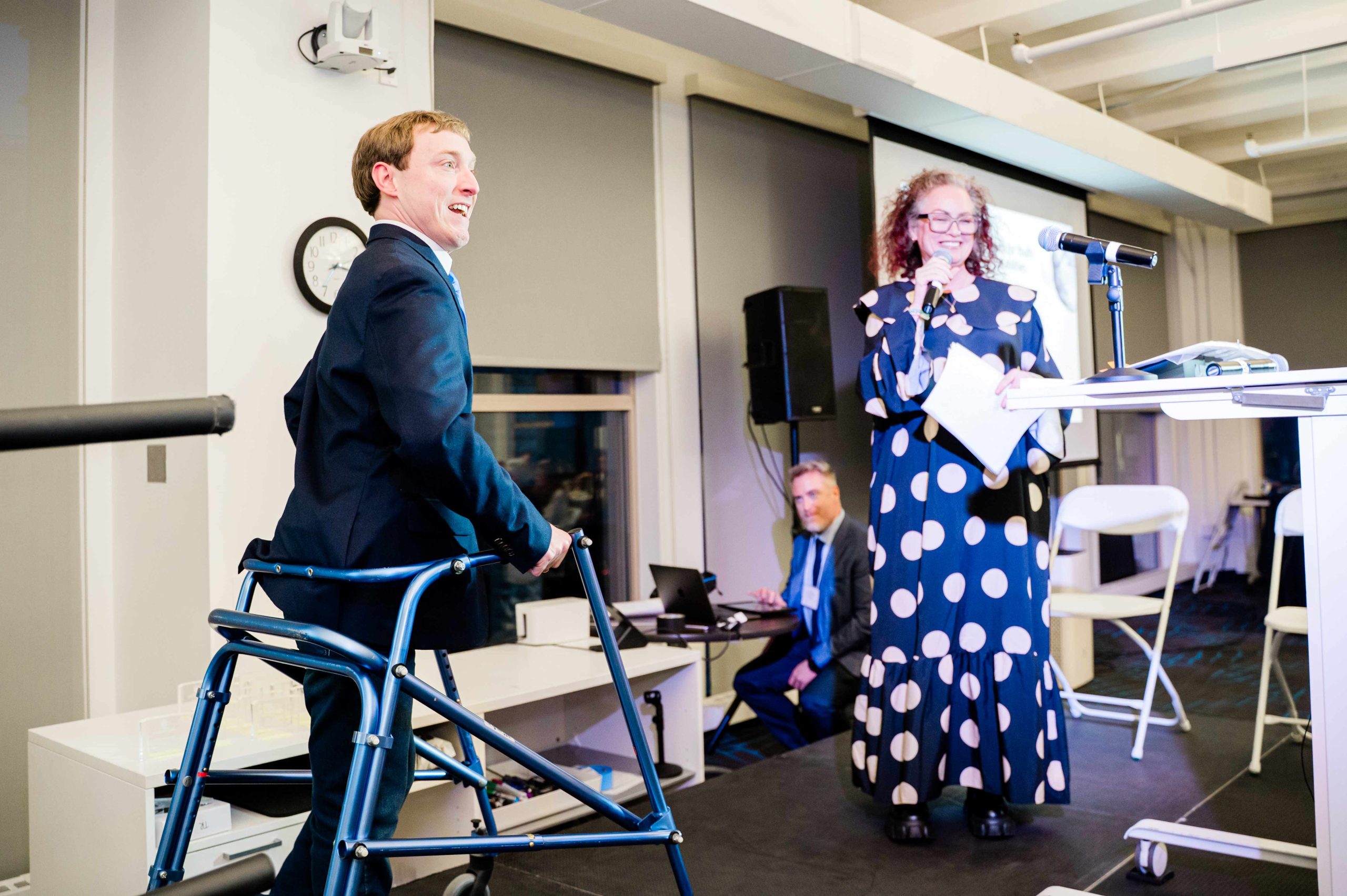 Austin using his walker to go on stage, as Kennedy holds a microphone