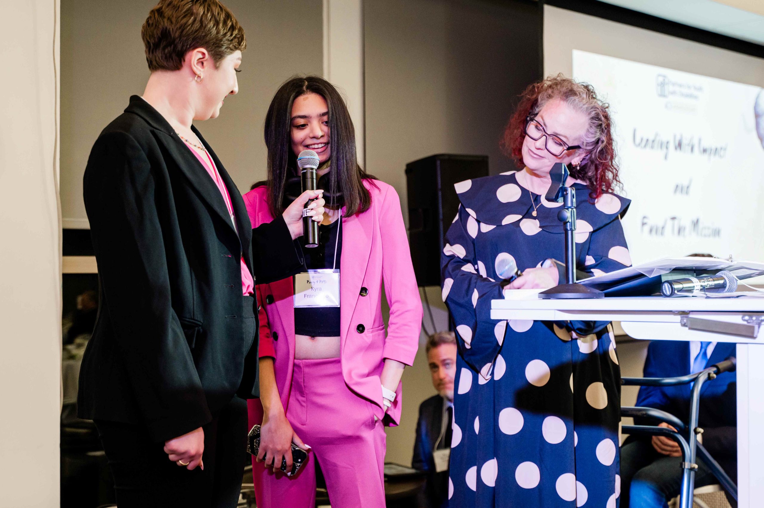 Mentor and Mentee duo, Sarah and Kyra are speaking into a microphone with Kennedy on the right.