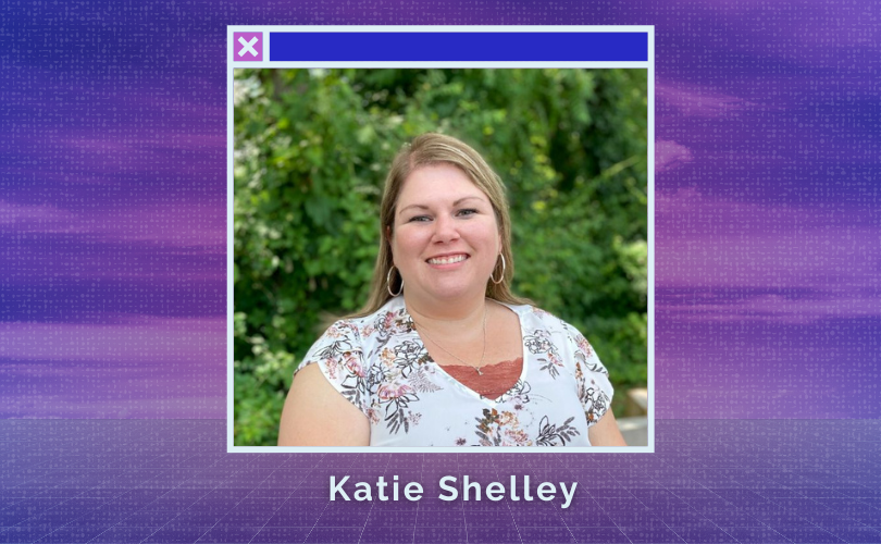 The head and shoulders of a white woman with long blonde hair, silver hoop earrings, wearing a white top with a floral motif. She smiles against a backdrop of green foliage.