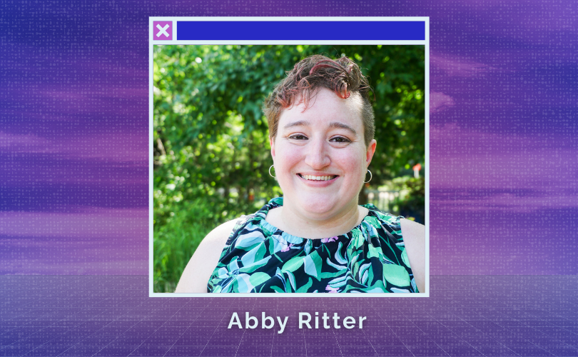 Abby, a white person with brown eyes and short curly hair, smiles at the camera. She is wearing a green floral dress and small hoop earrings.