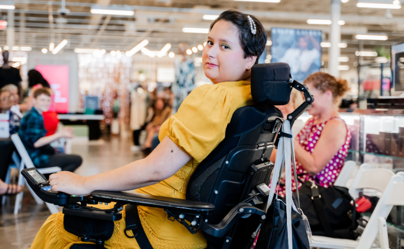 A PYD participant in a wheelchair, wears a yellow dress; she looks at the camera.