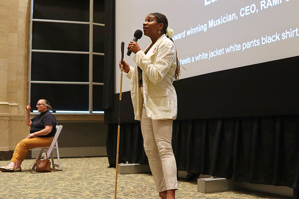 Singer, Lachi, standing, speaks into a microphone in front of a giant screen. An sign language interpreter sits far off to the side.