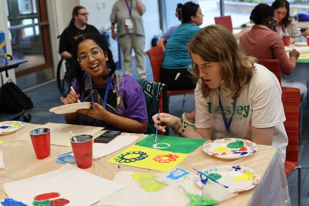 Participants sit at a table, painting with a variety of acrylic paint colors.